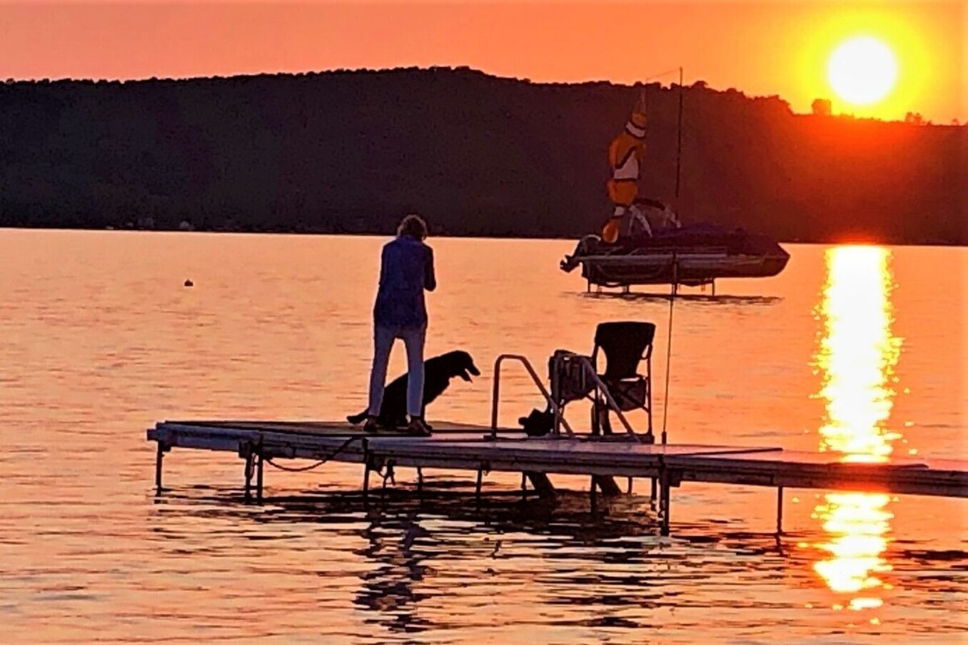 Een zonsondergang boven een rustig wateroppervlak met een steiger, een persoon en een hond.