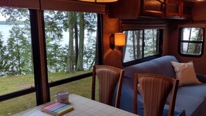 A cozy RV interior with a sofa, chairs, a table with a mug, and a view of a lake and trees. - Twin Birch Resort - The Loon on the Lake - Lakefront with Dock, Kayak, and Rowboat! (Honor)
