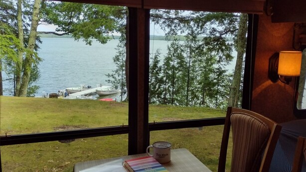 A view from a room with large windows overlooking a lake, a dock with boats, and a grassy area with trees. - Twin Birch Resort - The Loon on the Lake - Lakefront with Dock, Kayak, and Rowboat! (Honor)