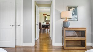 A hallway with a wooden floor leading to a dining area with a table and chairs. There is a lamp on a nightstand and a framed quote on the wall. - Charming Rancher, Close to Everything! (Joplin)