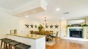 A modern kitchen with a central island, bar stools, a dining area with chairs, a fireplace, and decorative star wall art. - Game Room! Group Getaway 16 Mi to Galveston (Hitchcock)