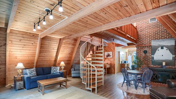 A cozy loft-style room with a wooden ceiling, a spiral staircase, a blue sofa, a dining table with chairs, and a rustic brick wall.