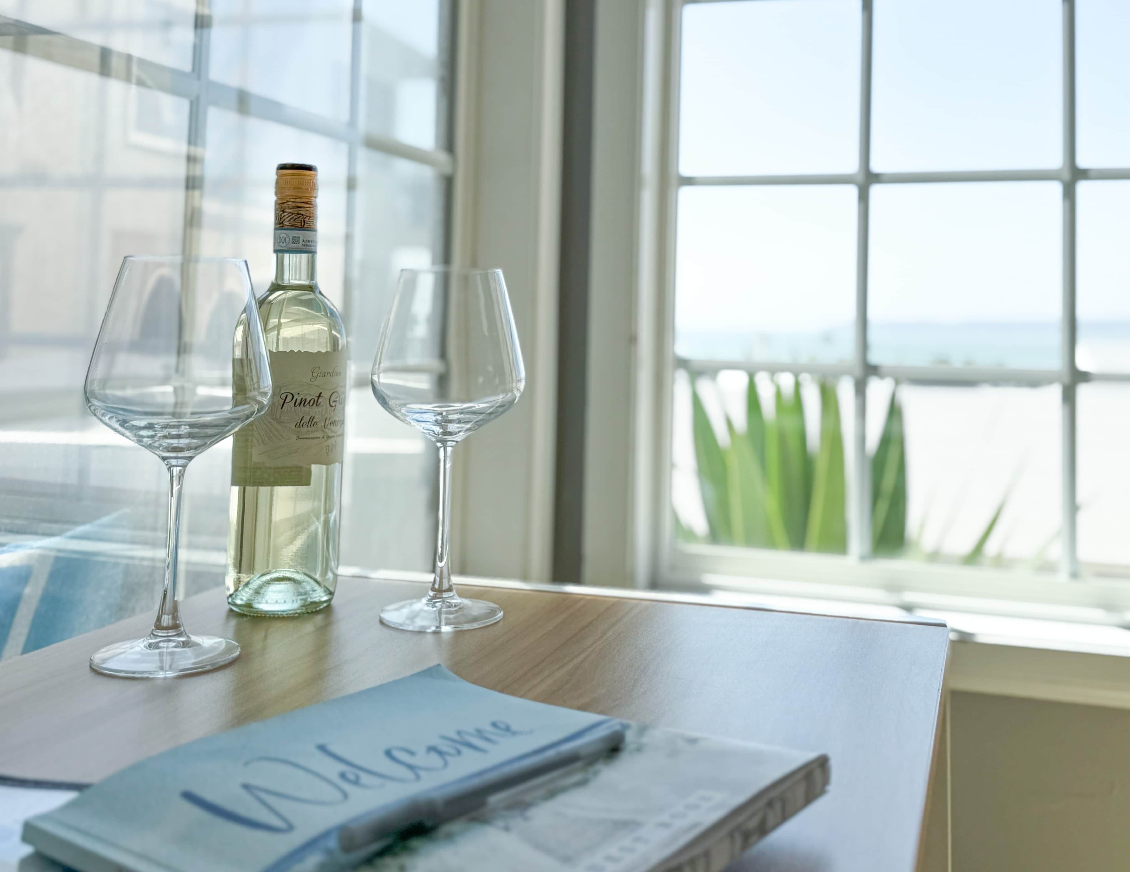 Ein Tisch mit einer Flasche Pinot Grigio und zwei leeren Weingläsern, ein Notizbuch mit dem Schriftzug „Welkommen“ und Blick auf das Meer durch ein Fenster.
