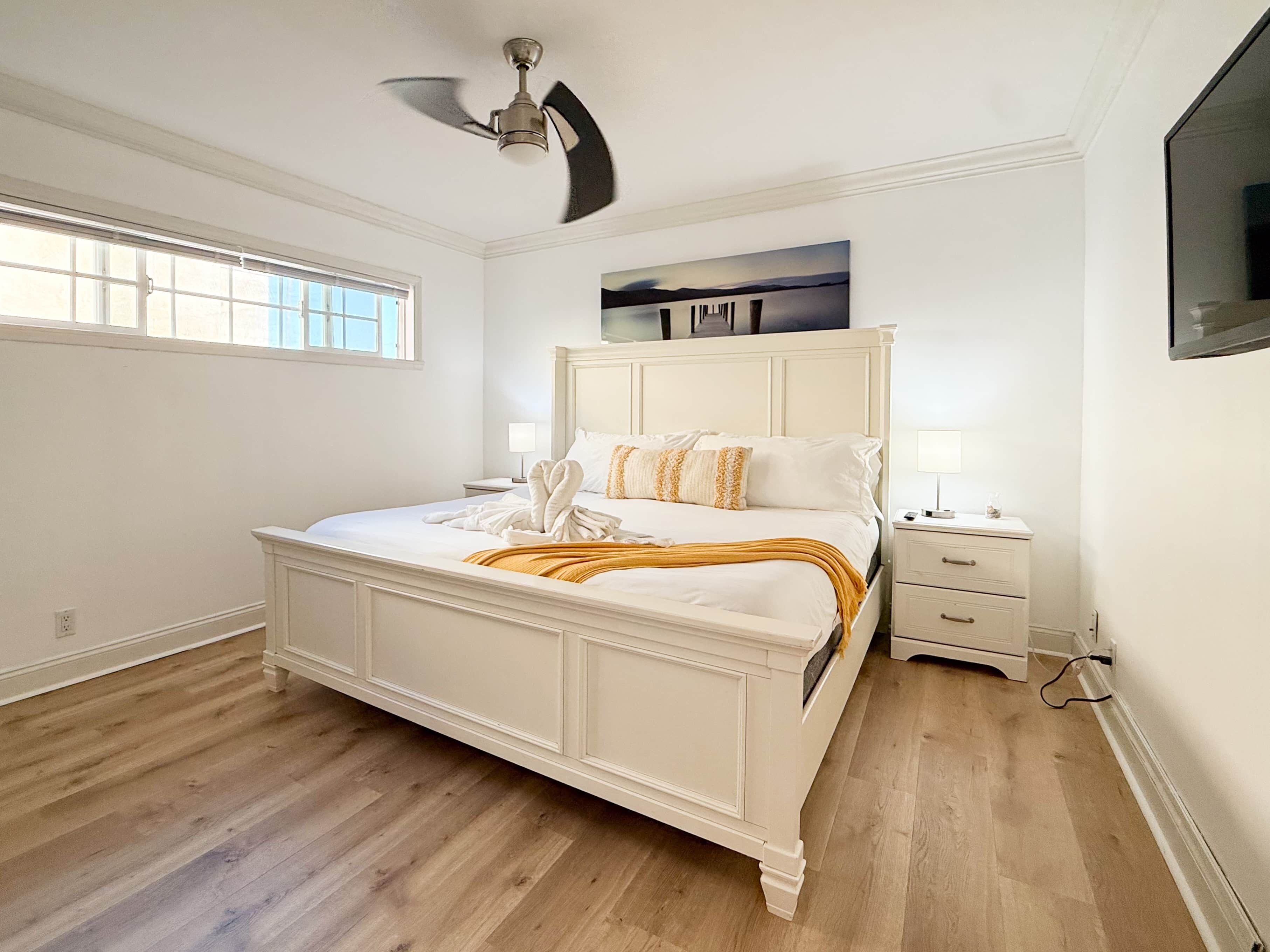 A bedroom with a large bed, a ceiling fan, a nightstand, a TV, and a window.