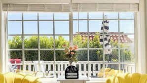 A bright room with a large window, two yellow chairs, a small table with a vase of flowers, and a white fence visible through the window. - Golden Coast Ocean View Penthouse (San Clemente)