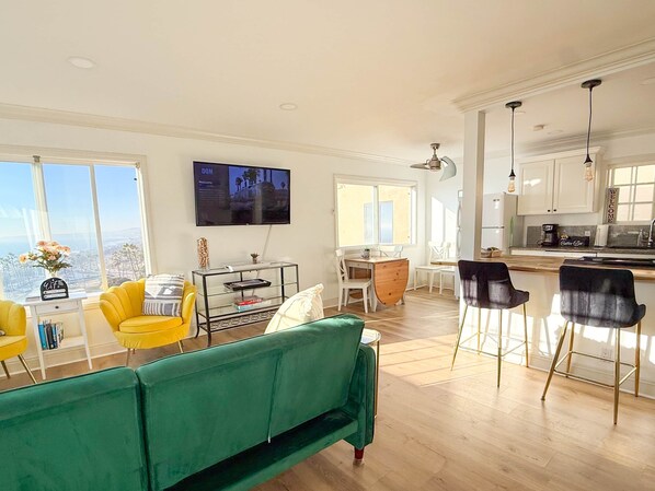 Una sala de estar moderna con un sofá verde, sillas amarillas y una zona de comedor con una mesa y sillas de madera. Un televisor está montado en la pared, y hay una cocina con taburetes de bar.