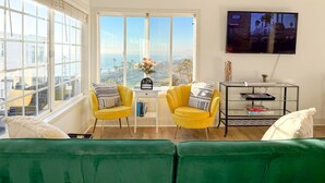 A living room with a large window, two yellow chairs, a green sofa, a TV on the wall, and a small table with books and a vase of flowers. - Golden Coast Ocean View Penthouse (San Clemente)