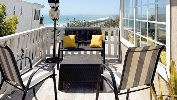 A balcony with a black wicker table and chairs, a striped umbrella, and a view of the ocean. - Golden Coast Ocean View Penthouse (San Clemente)