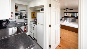 A compact kitchen with a gas stove, microwave, and a coffee machine. A small dining area is visible with a table and chairs. The room features a wooden floor and a bed with a headboard and pillows. - Ocean Bluff Hideaway (San Clemente)
