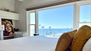 Une chambre avec un grand lit, un tableau accroché au mur et une vue sur la plage et l’océan par les fenêtres.
