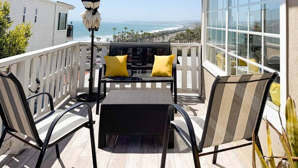 Un balcon avec une table en osier noir, deux chaises et un parasol à rayures.
