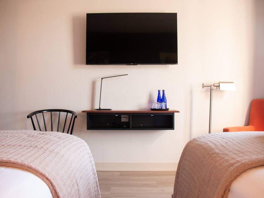 A hotel room with two beds, a TV mounted on the wall, a desk with a lamp, and a chair.