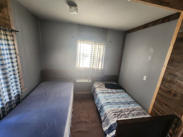 A small, compact room with two single beds, a window with curtains, and a wooden wall. - HUNTERS!! Are you looking for a house while you are in North Dakota? Here it is! (Tolna)