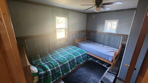 Una habitación pequeña con cama individual, cabecera de madera, ventilador de techo y dos ventanas que ofrecen luz natural.