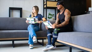 Zwei Personen sitzen auf einem grauen Sofa mit schwarzem Rahmen und lesen Zeitschriften.