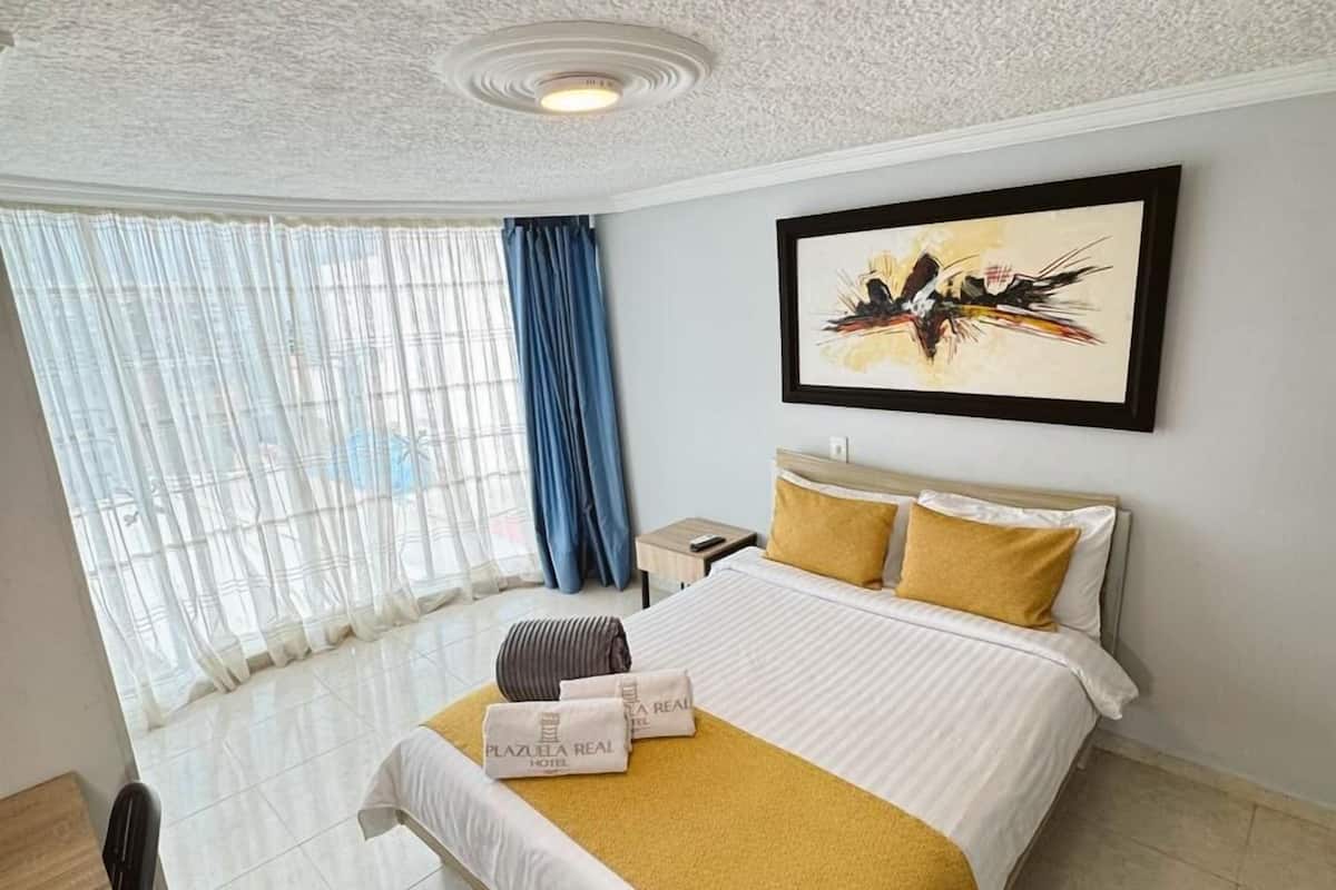 A hotel room with a bed, a desk, a chair, a nightstand, and a window with curtains.