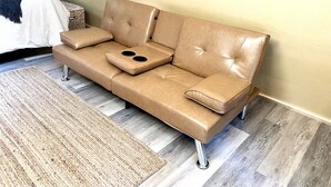 A tan leather sofa with armrests and a footrest, positioned against a wall with a wooden floor. - The EH Frame with Tree Pod in the woods! (Fountain Run)
