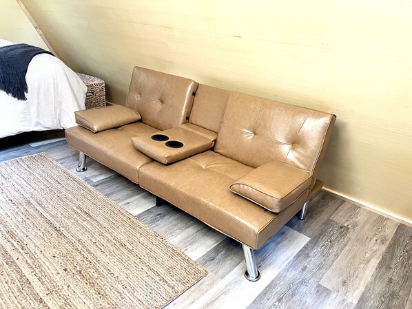 A tan leather sofa with armrests and a footrest, positioned against a wall with a wooden floor. - The EH Frame with Tree Pod in the woods! (Fountain Run)