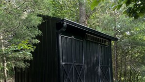 A black metal shed with a wooden door and a small step leading up to it, surrounded by trees and gravel. - The EH Frame with Tree Pod in the woods! (Fountain Run)