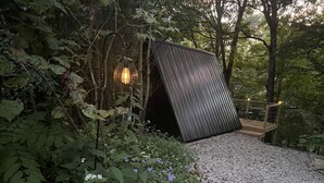 A wooden deck with a metal railing, surrounded by dense foliage and a gravel path. - The EH Frame with Tree Pod in the woods! (Fountain Run)