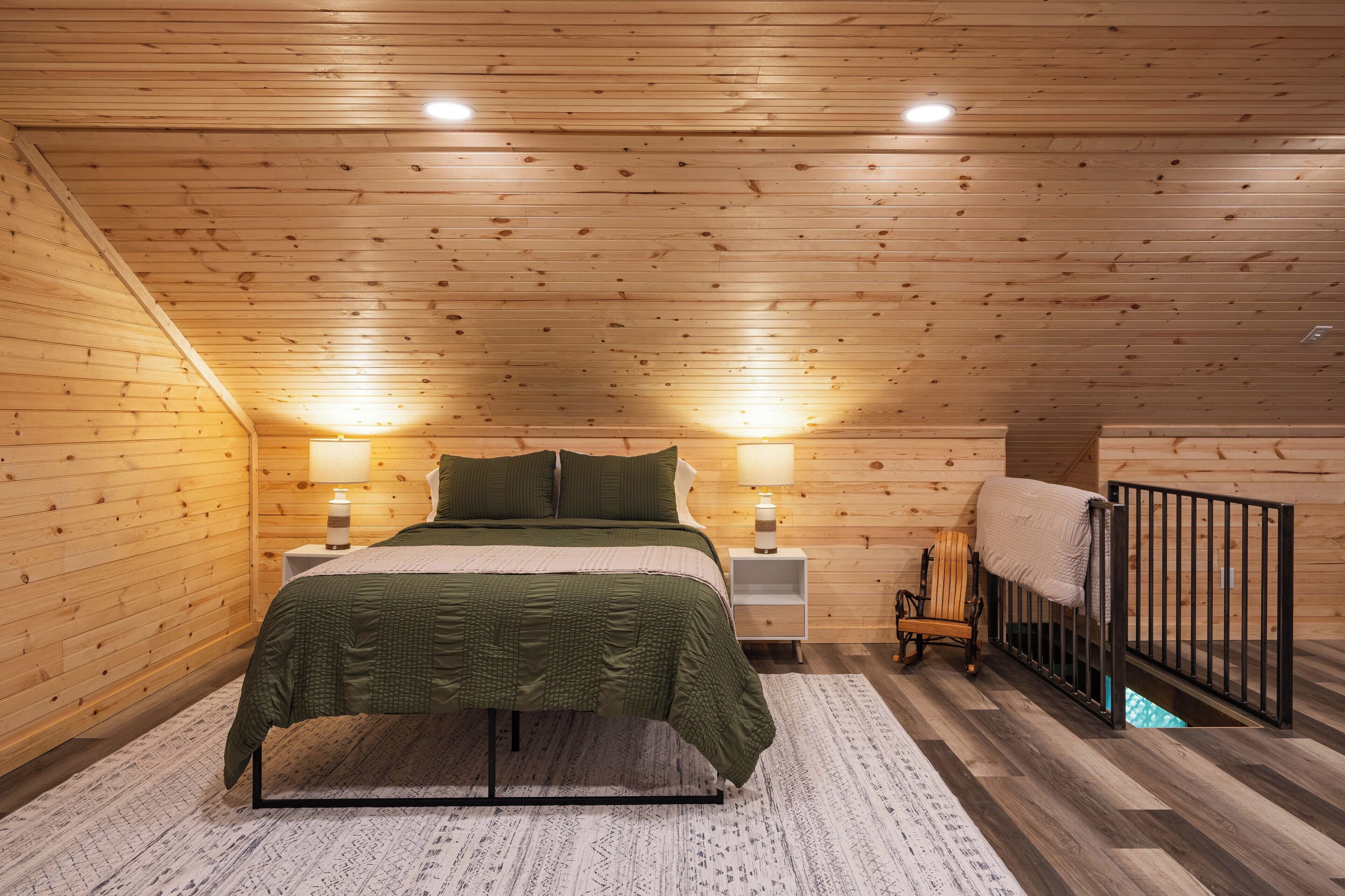 Ein Schlafzimmer mit Holzbalkendecke, einem Bett mit grüner Bettwäsche, zwei Nachttischlampen, einem Nachttisch, einem Sessel und einem Babybett.