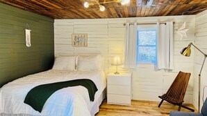 A bedroom with a wooden ceiling, a bed with white bedding, a nightstand with a lamp, a window with curtains, and a chair. - Rustic Old Mill Tiny Home (Fountain Run)