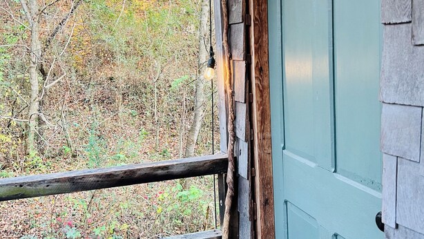 A wooden cabin with a teal door, a wooden railing, and a view of a forested area. - Rustic Old Mill Tiny Home (Fountain Run)