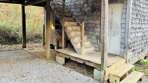 A rustic wooden staircase leading to a door, surrounded by a wooden exterior wall and a gravel ground. - Rustic Old Mill Tiny Home (Fountain Run)