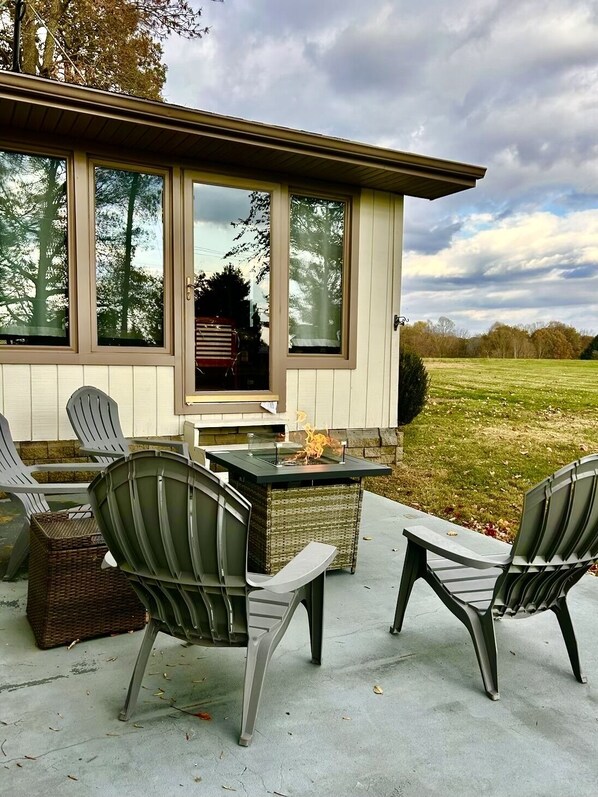 A patio with Adirondack chairs, a fire feature, and a view of a grassy field.