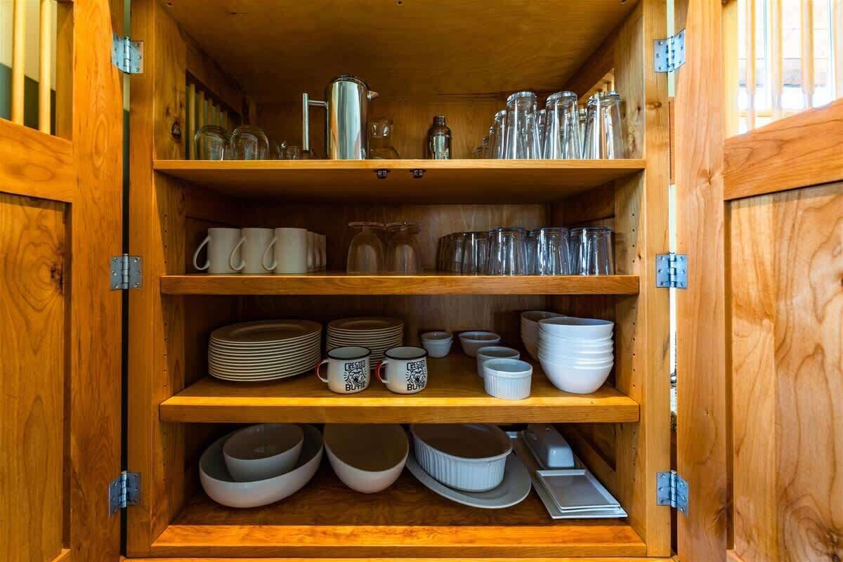 Un armadietto in legno con ripiani che contiene stoviglie da cucina, tra cui tazze, piatti, ciotole e bicchieri.