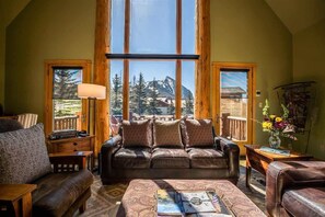 A cozy living room with a leather sofa, a coffee table with books, and a view of mountains through large windows.