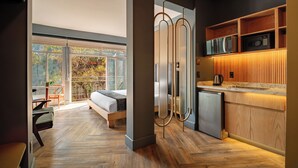Habitación de hotel moderna con suelo de madera, una cama, un escritorio, una silla, una zona de cocina con microondas y cafetera, y un ventanal con vistas a árboles.