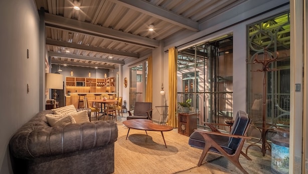 Una sala moderna con un sofá de cuero, una mesa de centro de madera y una silla azul. Se ve un área de cocina al fondo con armarios de madera y una mesa de comedor.