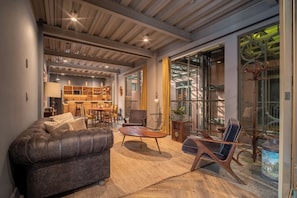 Un salon moderne avec un canapé en cuir, une table basse en bois et un fauteuil bleu. On aperçoit à l’arrière-plan un coin cuisine avec des meubles en bois et une table à manger.