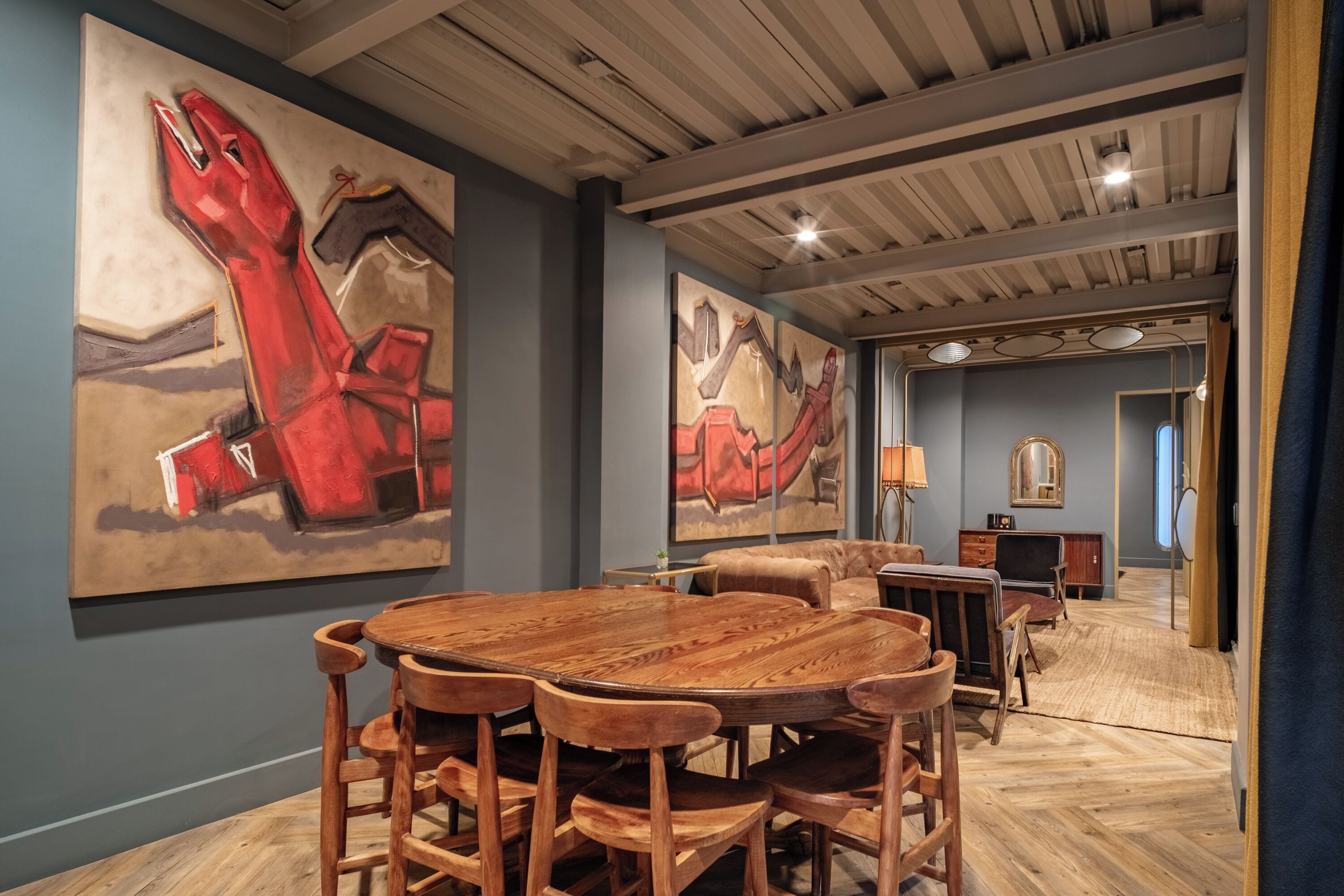 Un coin repas avec une table et des chaises en bois, de grandes peintures abstraites accrochées aux murs, et une vue sur une autre pièce où l’on aperçoit un canapé et un miroir.