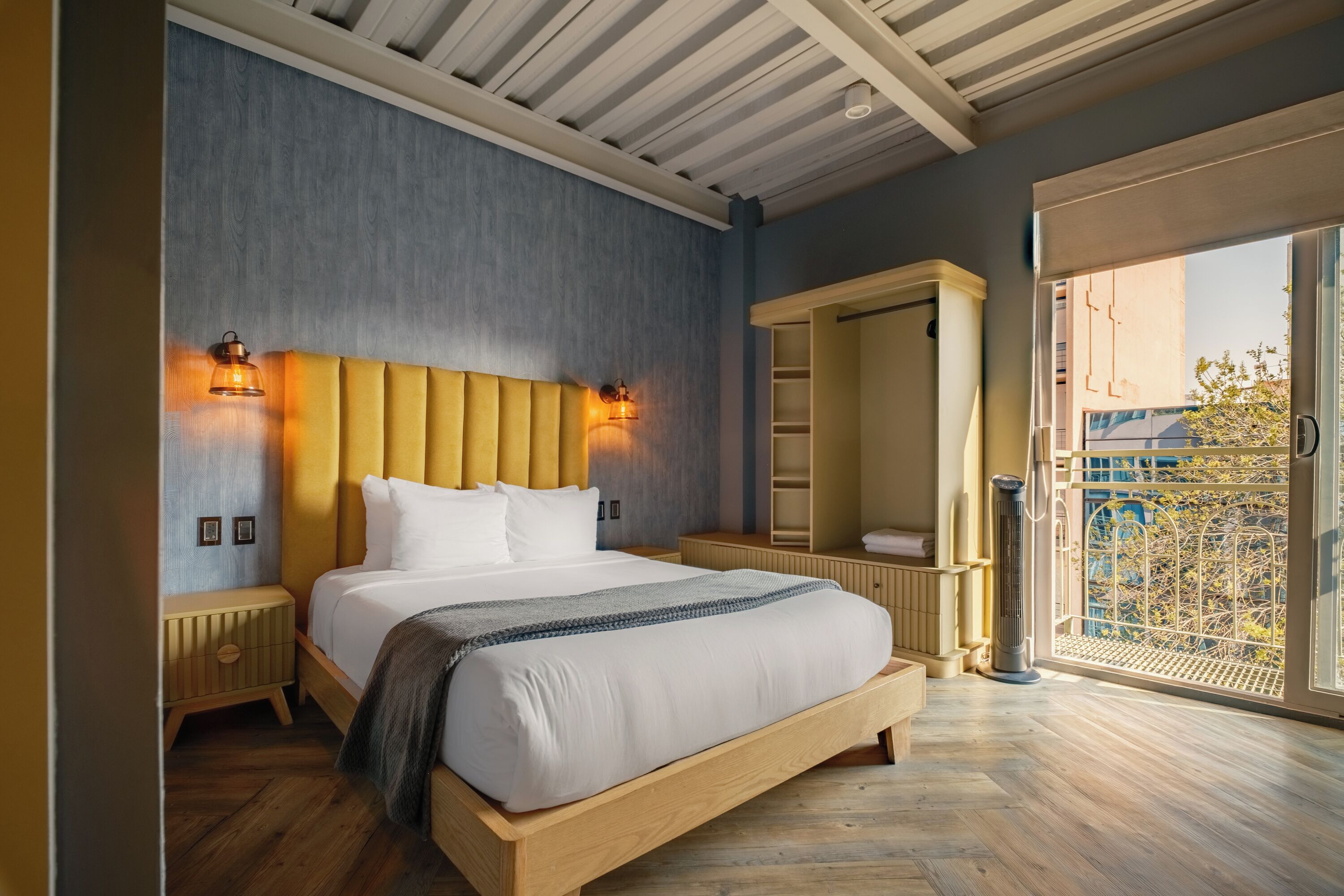 A modern bedroom with a large bed, wooden headboard, bedside lamps, a walk-in closet, and a large window with a view of the outdoors.
