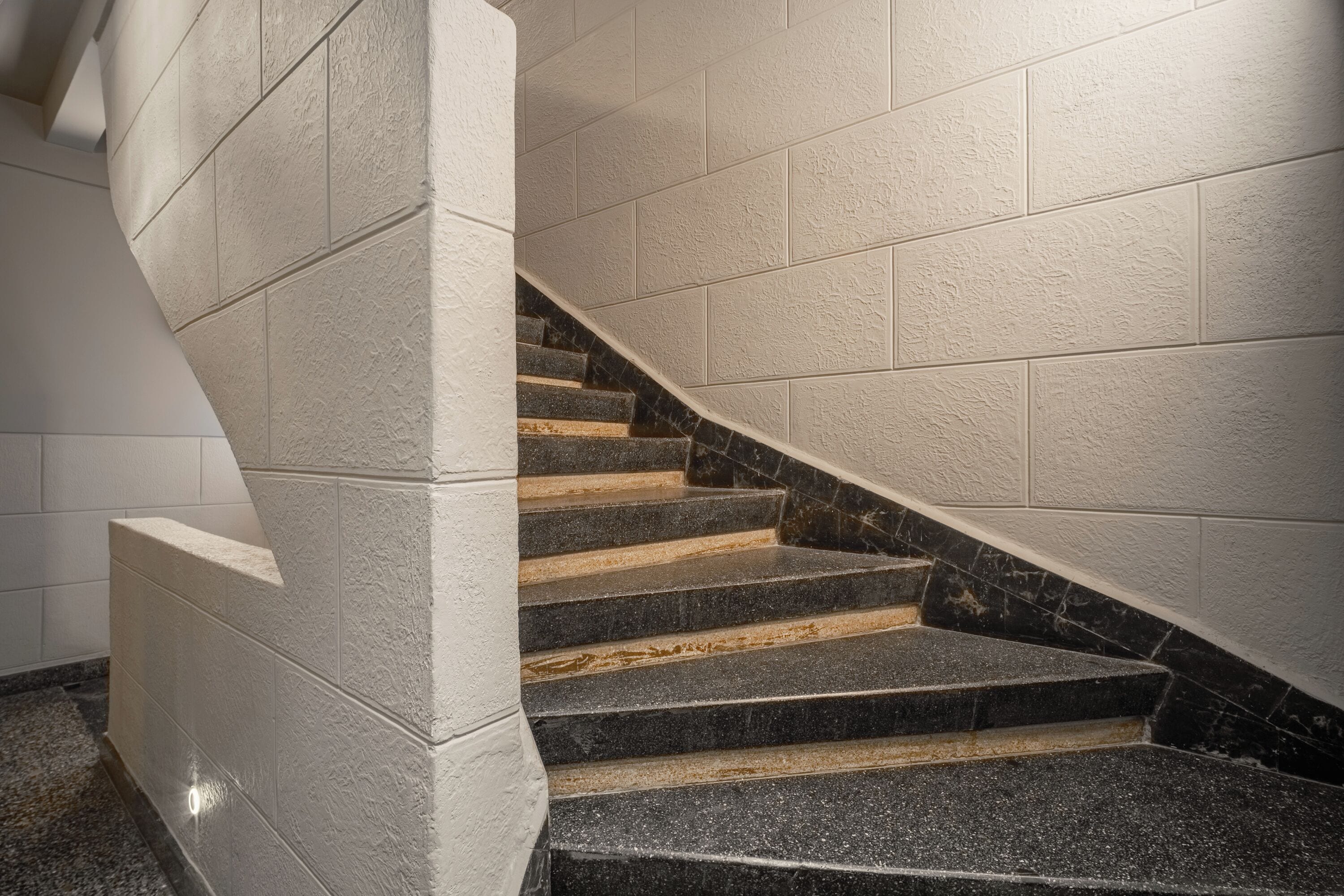 A spiral staircase with black and beige steps, white walls, and a granite floor.
