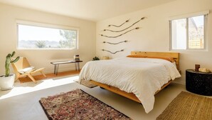 A bedroom with a large bed, a chair, a desk, a cactus, and a wall-mounted branch decoration.