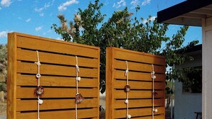 Two wooden chairs with slatted seats and backs, positioned in front of a wooden fence with horizontal slats and hanging decorative elements.