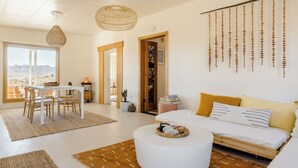 A modern living room with a white sofa, a dining table with chairs, a hanging light fixture, and a view of the desert through large windows.