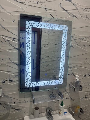 Un baño con pared de diseño de mármol, espejo con marco de vidrio, lavabo con grifo y dispensador de jabón.