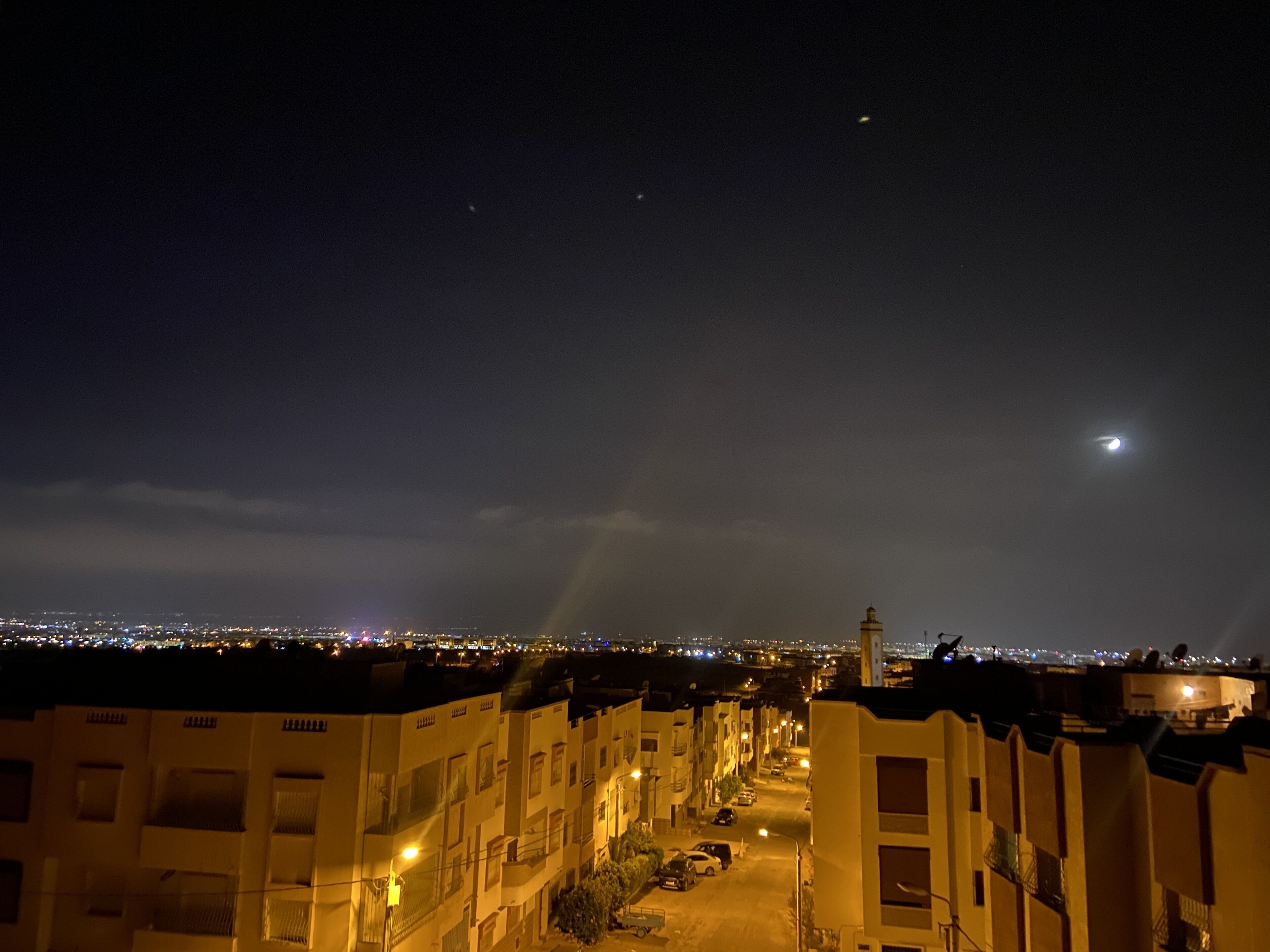 A nighttime view of a cityscape with illuminated buildings and a crescent moon in the sky.