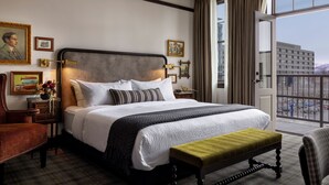 A hotel room with a large bed, a bench, a chair, a nightstand with a lamp, and a view of the city through the window. - The Avery Hotel (Boise)