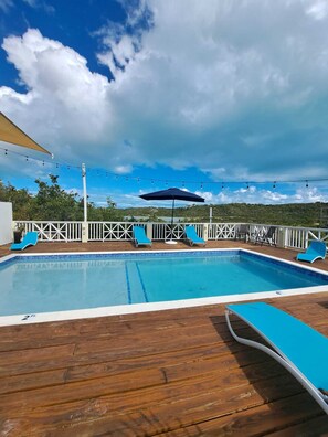 A rectangular pool with a wooden deck, surrounded by white railings and blue lounge chairs.