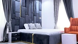 A modern bedroom with a large bed, a nightstand, a chair, and a decorative wall panel. - Spacious 3 bedroom  Apartment,  All room ensuite , Ipaja Lagos (Lagos)