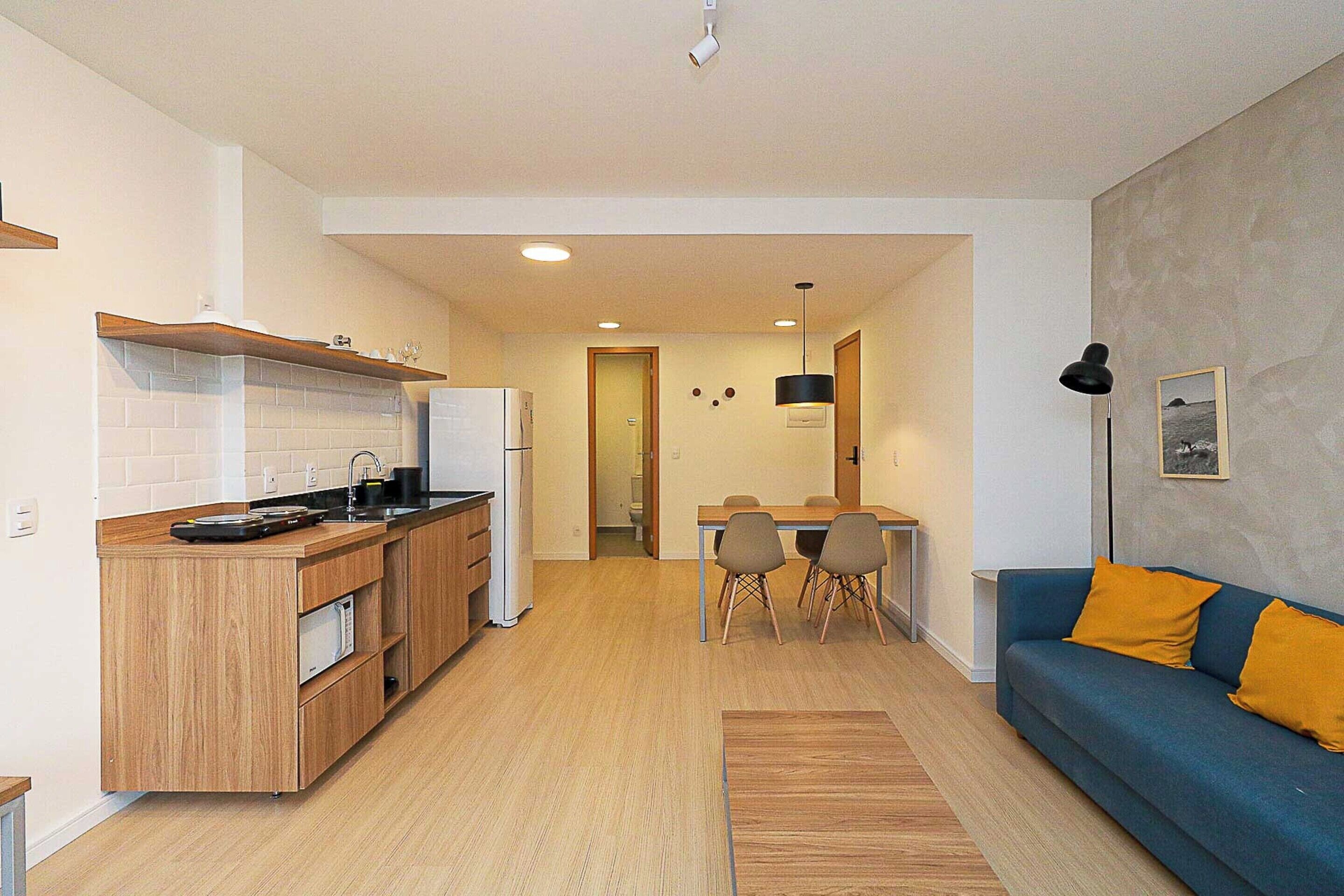A modern kitchen with wooden cabinets, a built-in oven, and a refrigerator. There is a dining area with a table and chairs, and a blue sofa with yellow cushions.