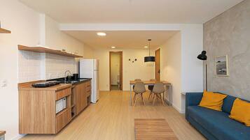 A modern kitchen with wooden cabinets, a built-in oven, and a refrigerator. There is a dining area with a table and chairs, and a blue sofa with yellow cushions.