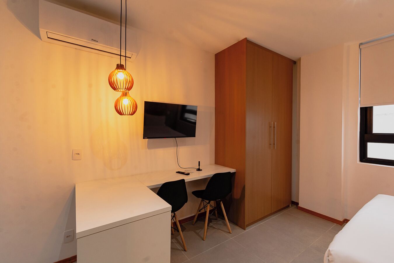 A modern room with a white desk, a black chair, a TV mounted on the wall, and a wooden wardrobe.