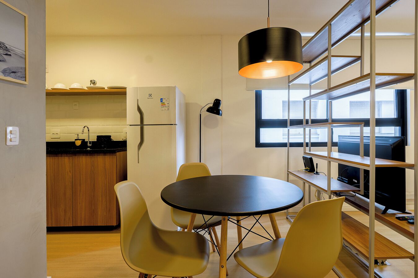 Cocina compacta con refrigerador, pileta y muebles de madera. Se observa una mesa de comedor con sillas e iluminación colgante.