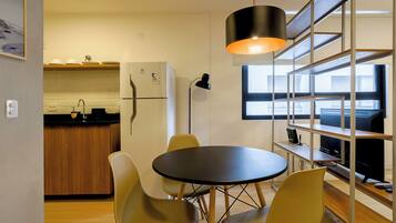 Cocina compacta con refrigerador, pileta y muebles de madera. Se observa una mesa de comedor con sillas e iluminación colgante.